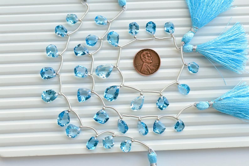 May include: A strand of light blue topaz gemstone beads strung on silver wire with silver bead accents. The beads are faceted and have a teardrop shape. The strand is finished with blue tassel charms.