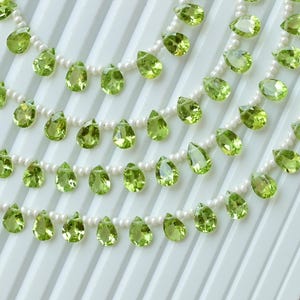 Puede incluir: Varias hebras de collares con piedras preciosas de peridoto verde claro en forma de pera. Las piedras de peridoto están ensartadas en delicadas hebras de perlas blancas, creando un efecto en capas. El fondo es una superficie blanca y acanalada.