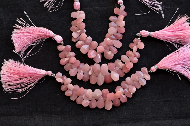 May include: Three strands of pink gemstone beads with tassels on each end. The beads are faceted and have a light pink color.