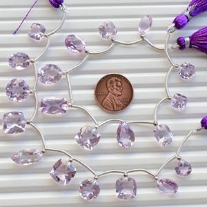 May include: A strand of pale pink amethyst gemstone beads strung on a silver chain with two purple tassels. The beads are various shapes and sizes, including oval, rectangular, and square. A UK penny is included for scale.