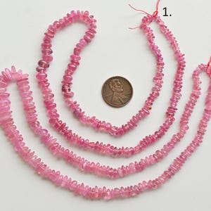 May include: Three strands of pink tourmaline beads, each strand is a different length. The beads are faceted and have a slightly irregular shape. The strands are laid out on a white surface with a US penny for scale.