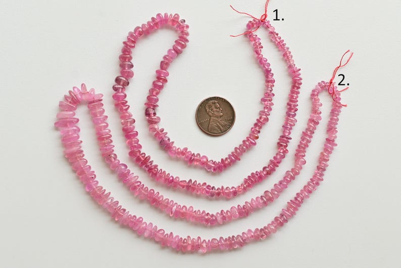 May include: Three strands of pink tourmaline beads, each strand is a different length. The beads are faceted and have a slightly irregular shape. The strands are laid out on a white surface with a US penny for scale.