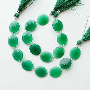 May include: A strand of faceted, circular green gemstone beads arranged in a loose coil. The strand is finished with two dark green decorative tassels attached to the ends with silver-toned metal findings, creating a cohesive jewellery component.