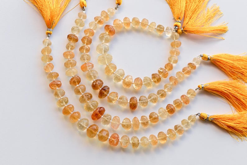 May include: A strand of golden citrine gemstone beads with a faceted, teardrop shape. The beads are strung on a cord with a tassel at each end. The tassels are a bright yellow color.