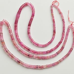 May include: Three strands of pink tourmaline beads, each strand is made of small, square-shaped beads. The beads are strung on a red thread.