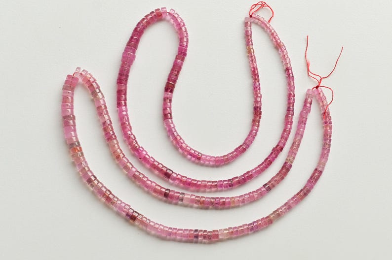 May include: Three strands of pink tourmaline beads, each strand is made of small, square-shaped beads. The beads are strung on a red thread.