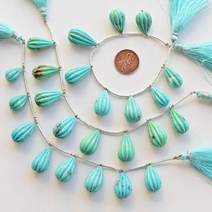 May include: A strand of turquoise gemstone beads with a teardrop shape. The beads are strung on silver wire and have a carved, fluted design. The strand is accented with turquoise tassels.