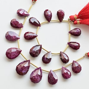 May include: A strand of 18 faceted pear-shaped ruby gemstones, each with a gold-toned bail, strung on a gold-toned wire. The strand is displayed on a white surface with red tassels on either end.