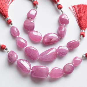 Może przedstawiać: Trzy sznury różowych koralików z kamieni szlachetnych z czerwonymi frędzlami na każdym końcu. Koraliki są gładkie i owalne.
