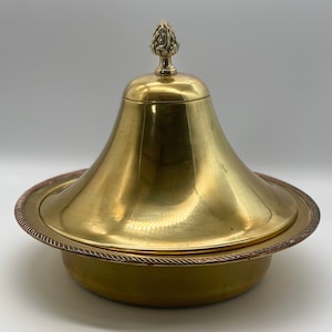 May include: A gold-coloured metal serving dish with a lid. The lid has a conical shape and is topped with a decorative finial. The dish has a round base with a decorative rope-like border. The overall design is ornate and vintage.