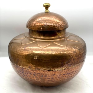 May include: A copper-coloured, hammered metal jar with a lid. The jar has a round body and a domed lid with a small, gold-coloured knob. The surface of the jar and lid is textured with small, evenly spaced indentations.