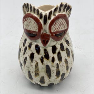 May include: A ceramic owl ornament with a cream-coloured body speckled with dark brown spots. The owl has large, reddish-brown eyes and a small, reddish-brown beak. The top of the owl's head and ears are detailed with dark brown accents.