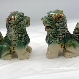 May include: A pair of green and white ceramic foo dog figurines. The foo dogs are in a seated position with their tails curled around their legs. They have intricate details and a glazed finish.