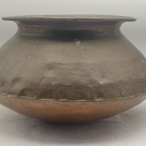 May include: A large, round, copper pot with a wide, flat base and a slightly flared rim. The pot has a dark brown patina and shows signs of age and use.