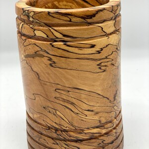 May include: A cylindrical wooden container with a natural wood grain pattern. The container has a light brown colour with dark brown streaks and horizontal grooves. The top and bottom edges are rounded. The container is approximately 15 cm tall.