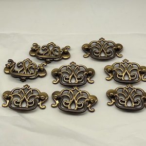 May include: A set of seven matching brass drawer pulls, each featuring an intricate, openwork scroll design with a central arched handle. The metal has a warm, aged patina with darker recessed areas, providing a consistent look for furniture hardware.