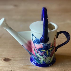 May include: A decorative ceramic watering can with a white spout and handle. The can features a blue handle and a floral design with blue, yellow, and green accents. The base and handle are a deep blue colour.