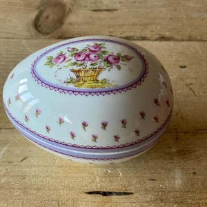 May include: A white porcelain egg-shaped trinket box with a floral design. The lid features a basket of pink roses with green leaves and a purple and white trim. The body of the box has a repeating pattern of small pink flowers.