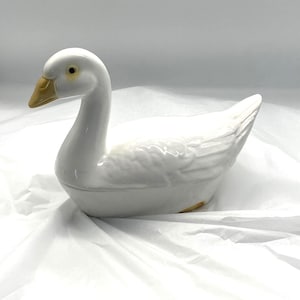 May include: A white ceramic goose ornament with a curved neck and a yellow beak. The goose has a detailed wing design and is resting on a white surface. The ornament is a decorative item.