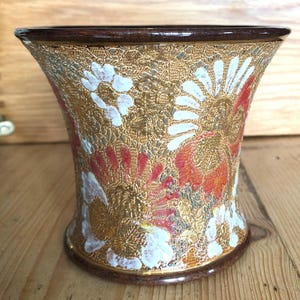 May include: A ceramic vase with a gold and brown glaze. The vase has a wide mouth and a narrow base. It is decorated with white and red floral patterns.