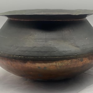 May include: A large, round copper pot with a dark patina. The pot has a wide, flat base and a rounded body. The copper is tarnished and has a greenish hue in some areas.