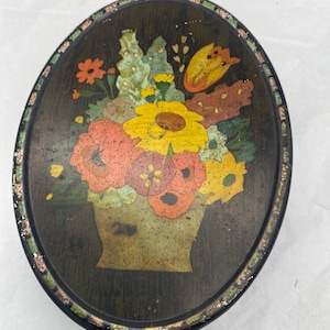 May include: An oval-shaped tin box with a floral design. The tin is painted with a dark brown background and features a bouquet of colourful flowers in a vase. The flowers are painted in shades of red, orange, yellow, and green.