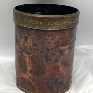 May include: A cylindrical copper vessel with a weathered, dark patina and a brass rim. The brass band is engraved with the text CASELLA LONDON No 9436. The copper body shows signs of oxidation and minor surface indentations throughout the exterior.