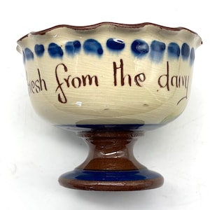 May include: A ceramic bowl with a pedestal base. The bowl is off-white with a scalloped rim and blue accents. The words "fresh from the dairy" are written on the side in brown script. The base is brown and blue.
