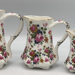 May include: Set of three white ceramic jugs with a floral pattern in shades of pink, yellow, and green. The jugs have a gold trim around the rim.
