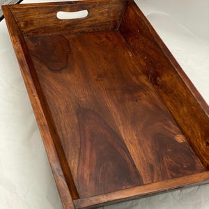 May include: A rectangular wooden serving tray with a dark brown finish and visible wood grain patterns. The tray features high sides and integrated cutout handles on the shorter ends, providing a sturdy structure for carrying items.
