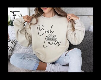 May include: A beige sweatshirt with the words "Book Lover" in a script font, with a floral book graphic. The person is wearing light blue jeans and white socks. The sweatshirt has long sleeves.