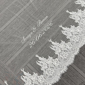 May include: A white wedding veil with a delicate lace trim. The veil has the names "Soraya & Roman" and the date "26/06/2026" embroidered in white script. The veil is made of a sheer fabric.