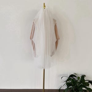 May include: A white tulle wedding veil displayed on a gold stand. The veil is single-layered and has a scalloped edge.