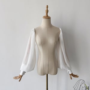 Könnte beinhalten: Eine weiße Bluse mit langen, transparenten Ärmeln und geknöpften Manschetten, präsentiert auf einer Schneiderpuppe. Die Ärmel haben eine leichte Puffärmel-Form an der Schulter und verjüngen sich zum Handgelenk. Die Schneiderpuppe hat eine neutrale beige Farbe.