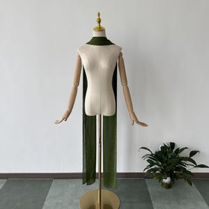 May include: A long, dark green scarf draped over a white mannequin. The scarf is made of a soft, flowing fabric and is long enough to be worn as a shawl or a wrap.