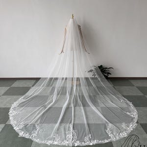 May include: A long, white bridal veil with a delicate floral lace trim along the edge. The veil is displayed on a mannequin, showcasing its length and flowing design. The veil is made of a sheer fabric, allowing light to pass through.