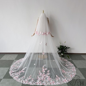 May include: A long, white bridal veil with delicate pink floral embroidery along the bottom and a band across the top. The veil is displayed on a mannequin, showcasing its flowing design and intricate details. The veil's train extends gracefully.
