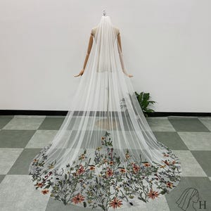 May include: A long, white bridal veil with embroidered flowers in red, orange, green, and yellow. The veil is displayed on a mannequin, highlighting its length and delicate details. The floral embroidery is concentrated at the bottom edge of the veil.