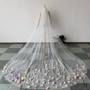 Puede incluir: Un velo de novia largo y blanco con intrincados bordados florales en tonos rosa, morado, amarillo y verde. El velo se exhibe en un maniquí, mostrando su delicado diseño fluido. Los detalles florales se concentran en el borde inferior.