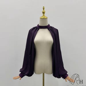 May include: A dark purple blouse with a high neckline and long, voluminous sleeves. The blouse is displayed on a mannequin, showcasing its open front design and elegant draping. The fabric appears to be a smooth, flowing material.
