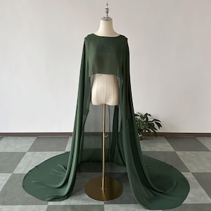 May include: A dark green cape with a flowing design, displayed on a mannequin. The cape features a round neckline and long, draping panels that extend to the floor. The fabric appears to be a lightweight, semi-sheer material.