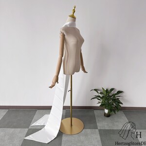 May include: A beige dress form with wooden arms and a golden head and base. A white fabric is draped over the form's shoulders and hangs down. The dress form is on a gold stand. The logo "HerzogStoreDE" is in the bottom right corner.