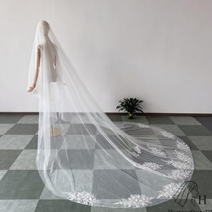 May include: A long, white bridal veil with delicate floral lace detailing along the edge. The veil is displayed on a mannequin, showcasing its flowing design. The veil's length extends across the floor, creating an elegant and romantic look.