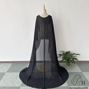 May include: A long, flowing black cape made of sheer fabric, displayed on a mannequin. The cape features a rounded neckline and a full, floor-length design. The fabric drapes elegantly, creating a dramatic effect. The cape is suitable for costumes or special events.
