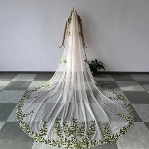 May include: A long, white tulle wedding veil with green floral embroidery. The veil is draped over a mannequin and has a scalloped edge.