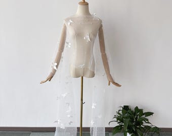 May include: A sheer white bridal veil with butterfly and pearl embellishments. The veil is draped over a mannequin, showcasing its delicate fabric and design. The butterflies are scattered across the veil, adding a whimsical touch to the bridal accessory.