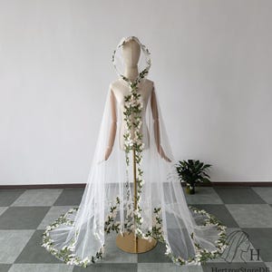 May include: A sheer white hooded cape with floral embroidery. The cape features a delicate design of white flowers and green leaves cascading down the front and along the hem. The cape is displayed on a mannequin.