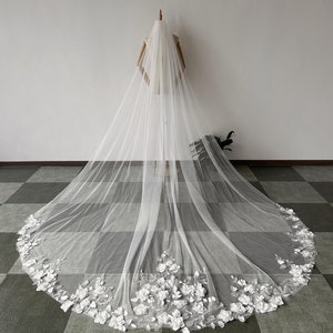 May include: A long white tulle wedding veil with a floral lace trim. The veil is draped over a mannequin and is shown from the back.