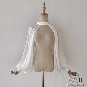 May include: A white satin blouse with long, voluminous sleeves and a high neckline. The blouse features a keyhole opening at the chest and is displayed on a mannequin. The sleeves gather at the wrists.