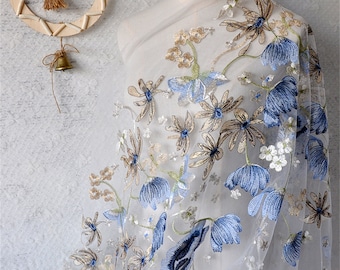 Puede incluir: Tela blanca transparente con diseño floral bordado. El bordado presenta flores azules, doradas y blancas con tallos verdes. La tela está drapeada sobre un maniquí, mostrando el delicado patrón floral.
